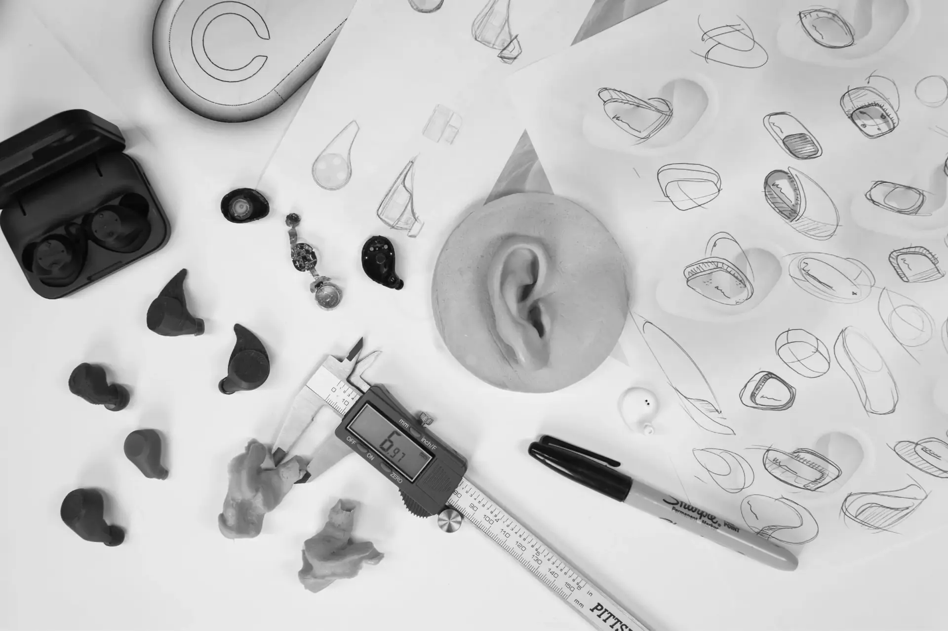 Earbud prototypes, a moulded ear model, calipers and concept sketches laid out on a desk